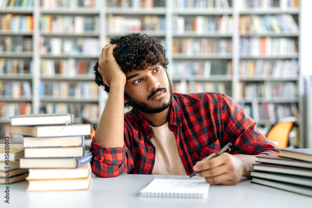 Sad arabian or indian male student tired from studying, sits at a desk in a library against the background of bookshelves, upset millennial guy looks away, feels exhausted from long reading books Sad arabian or indian male student tired from studying, sits at a desk in a library against the background of bookshelves, upset millennial guy looks away, feels exhausted from long reading books