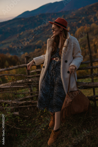 Fashionable woman wearing trendy autumn outfit with orange hat, glasses, faux fur coat, zebra print dress, holding trendy brown suede bag, posing in mountain landscape