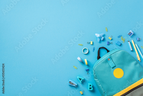 Back to school concept. Top view photo of blue rucksack plastic alphabet letters binder clips plane shaped sharpener eraser adhesive tape stapler and pens on isolated blue background with copyspace