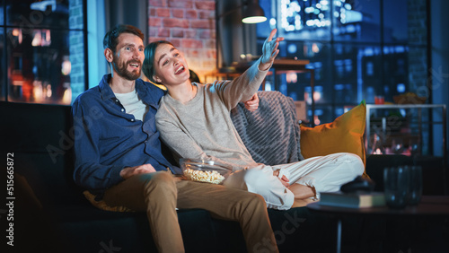 Portrait of Beautiful Couple Spending Time at Home, Sitting on a Couch, Hugging and Watching Exciting TV Show in Their Stylish Loft Apartment. Man and Woman Streaming Comedy Movie and Have Good Time. © Gorodenkoff