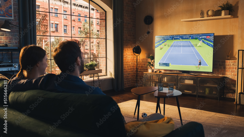 Authentic Couple Spending Time at Home, Sitting on a Couch and Watching Tennis Championship on Flat Screen Television Set. Man and Woman Enjoying Live Broadcast of Sports Finals. Authentic Couple Spending Time at Home, Sitting on a Couch and Watching Tennis Championship on Flat Screen Television Set. Man and Woman Enjoying Live Broadcast of Sports Finals.