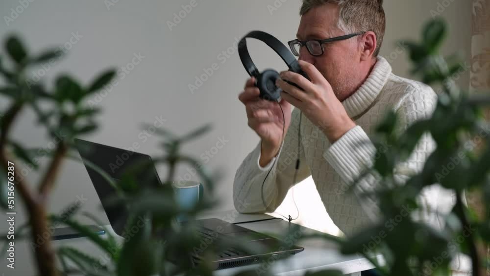 handsome mature adult man with glasses is sitting at a laptop in an office with green house plants in blurry form in the foreground. removes headphones and glasses to relax. sustainable environment.4k handsome mature adult man with glasses is sitting at a laptop in an office with green house plants in blurry form in the foreground. removes headphones and glasses to relax. sustainable environment.4k
