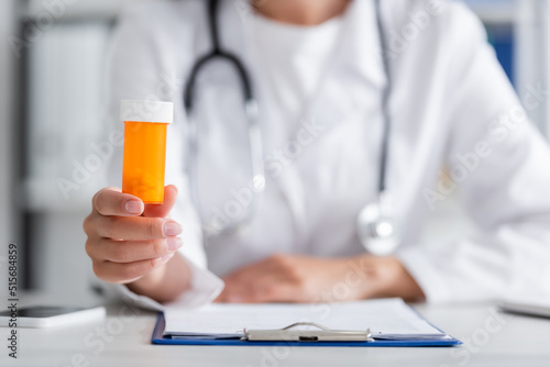 Cropped view of blurred doctor holding pills near clipboard and smartphone in clinic