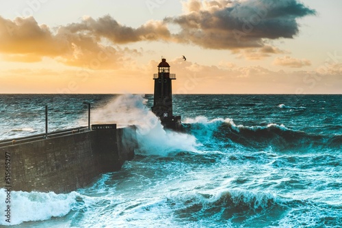 Large waves crash against the stone tower of the lighthouse at high tide at sunset © Wirestock Creators