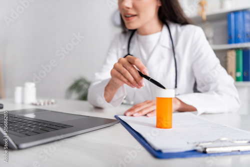 Cropped view of doctor pointing at jar with pills on clipboard during online consultation on laptop in clinic