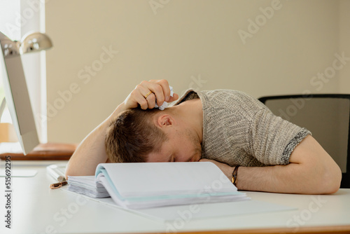 Depressed caucasian man with headache sitting at his desk in the office. Stressed young employee has problems at work. Businessman has burnout. Mental health. Overwhelmed by working. Deadline, stress.