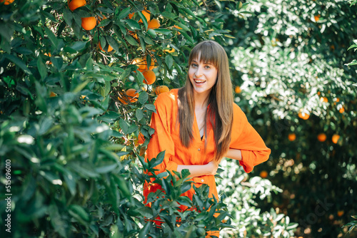Happy young girl in orange dress is posing to camera by putting hands together on waist in orange garden