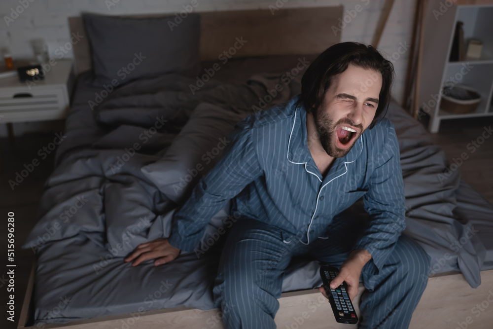 Tired man yawning and holding remote controller in bedroom at night