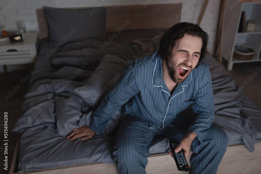 Tired man yawning and holding remote controller in bedroom at night Tired man yawning and holding remote controller in bedroom at night