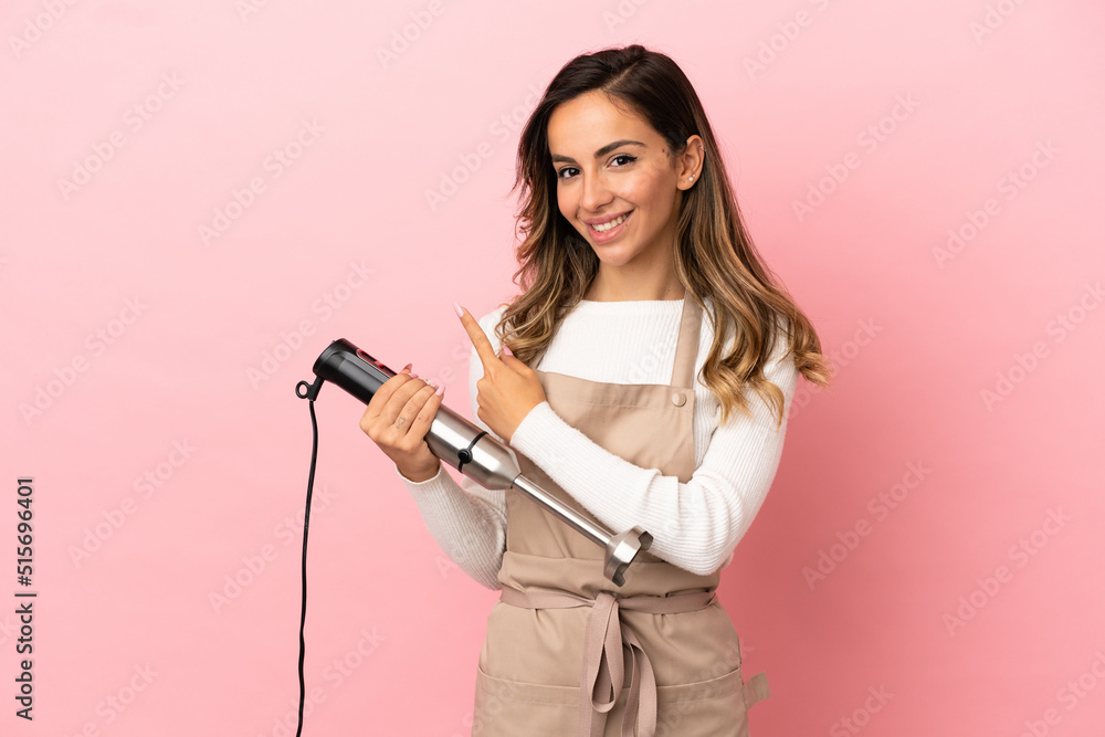 Young woman using hand blender over isolated pink background pointing to the side to present a product Young woman using hand blender over isolated pink background pointing to the side to present a product