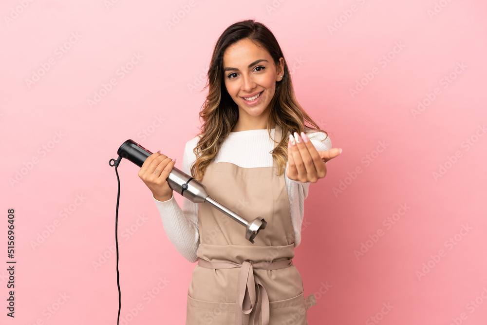 Young woman using hand blender over isolated pink background inviting to come with hand. Happy that you came Young woman using hand blender over isolated pink background inviting to come with hand. Happy that you came