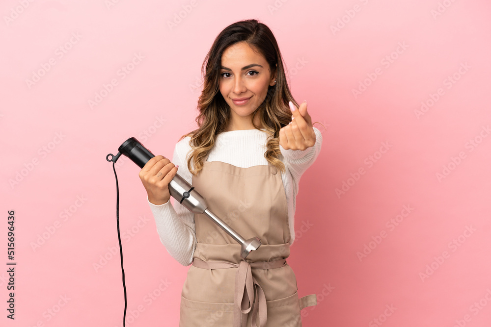 Young woman using hand blender over isolated pink background making money gesture Young woman using hand blender over isolated pink background making money gesture