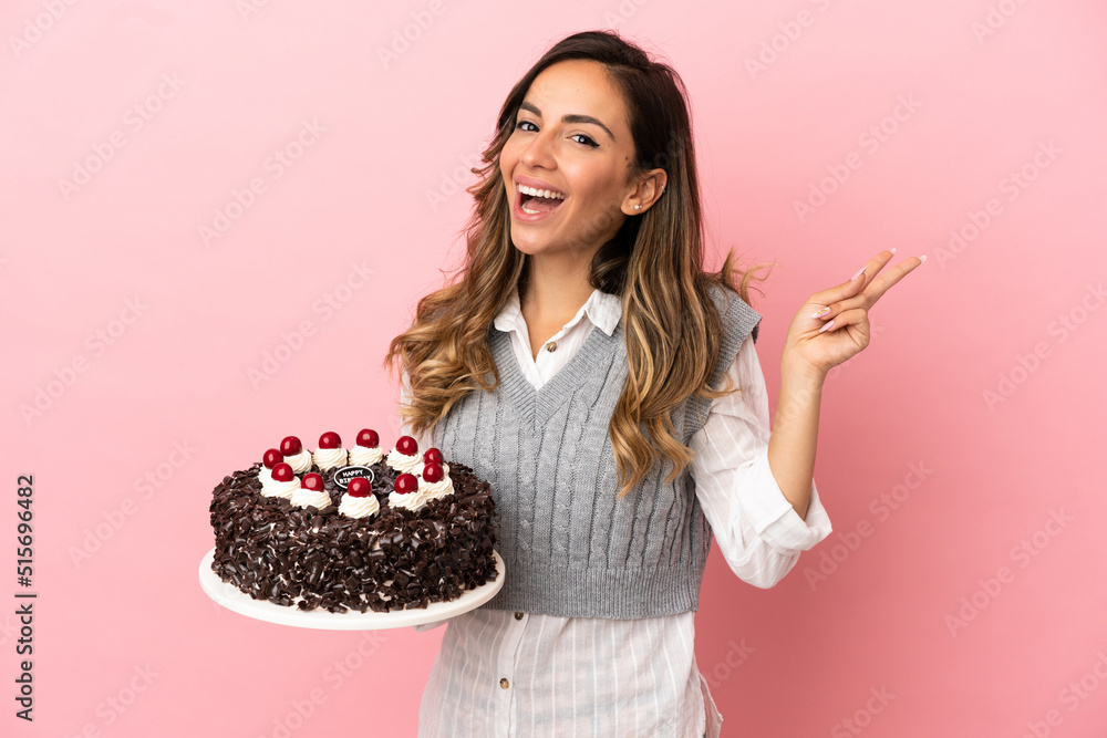 Young woman holding birthday cake over isolated pink background smiling and showing victory sign Young woman holding birthday cake over isolated pink background smiling and showing victory sign