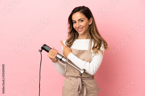 Young woman using hand blender over isolated pink background pointing to the side to present a product
