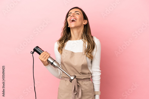 Young woman using hand blender over isolated pink background laughing