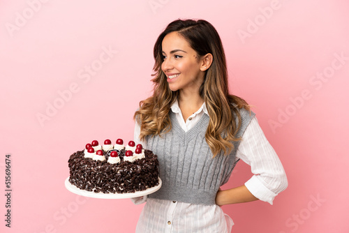 Young woman holding birthday cake over isolated pink background looking to the side and smiling
