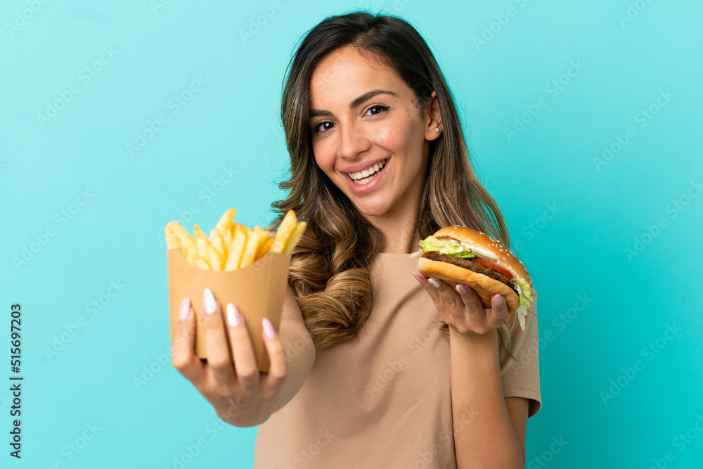 Young woman holding fried chips and burger over isolated background Young woman holding fried chips and burger over isolated background