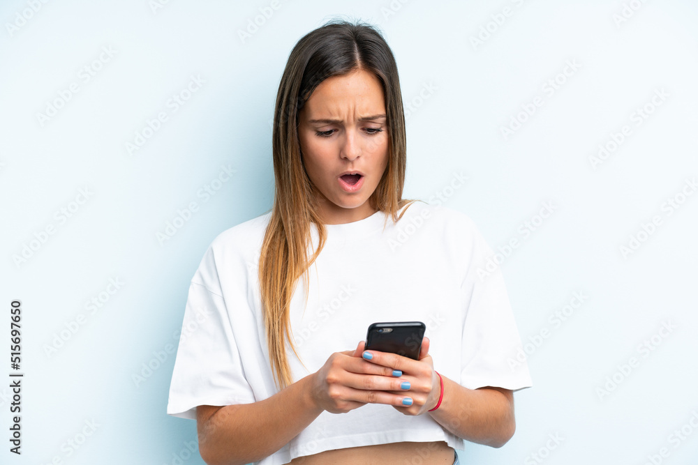 Young caucasian woman isolated on blue background looking at the camera while using the mobile with surprised expression Young caucasian woman isolated on blue background looking at the camera while using the mobile with surprised expression