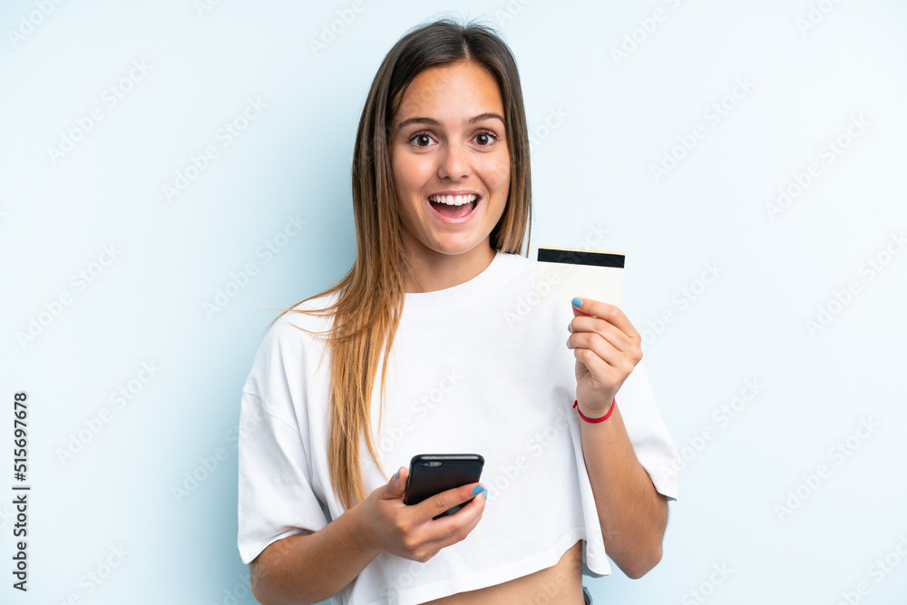 Young caucasian woman isolated on blue background buying with the mobile and holding a credit card with surprised expression Young caucasian woman isolated on blue background buying with the mobile and holding a credit card with surprised expression