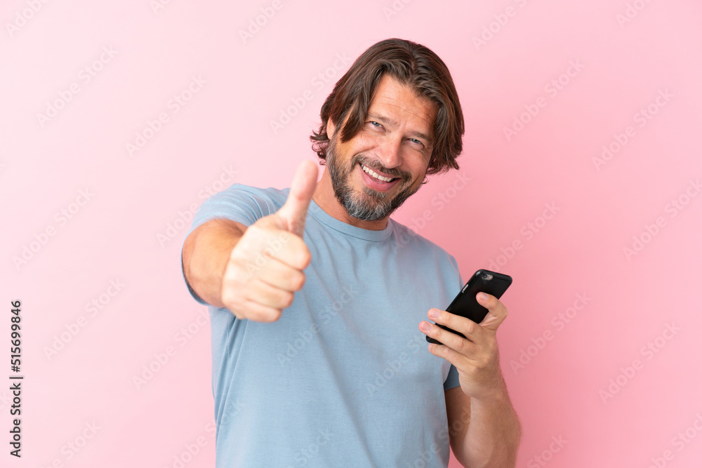 Senior dutch man isolated on pink background using mobile phone while doing thumbs up Senior dutch man isolated on pink background using mobile phone while doing thumbs up