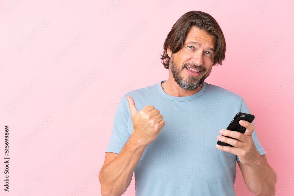 Senior dutch man isolated on pink background using mobile phone and pointing to the lateral Senior dutch man isolated on pink background using mobile phone and pointing to the lateral