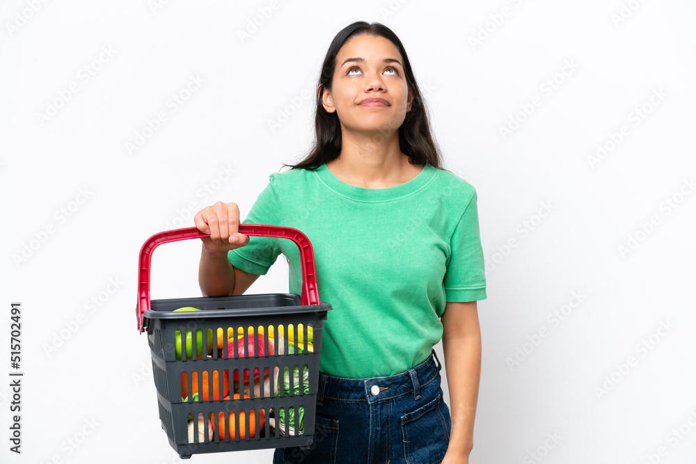 Young Colombian woman holding a shopping basket full of food isolated on white background and looking up Young Colombian woman holding a shopping basket full of food isolated on white background and looking up