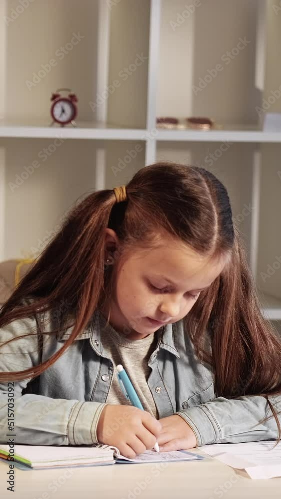 Vertical video. Kid homework. Primary school knowledge. Home learning. Diligent busy smart small girl studying writing exercise working at desk.