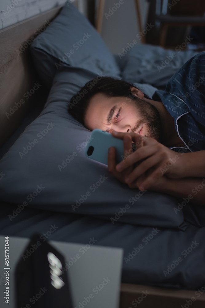 Sleepless man using mobile phone near blurred alarm clock on bed at night Sleepless man using mobile phone near blurred alarm clock on bed at night