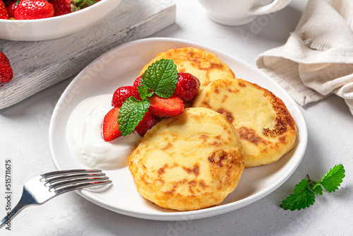 Cottage cheese pancakes with strawberries and cream in a white plate on a light gray background close-up. Side view.