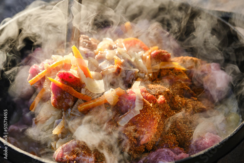 Cooking pilaf on a fire. Meat with carrot is cooking in cauldron on fire with smoke. Shallow depth of field