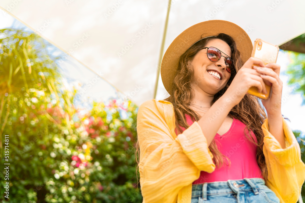 young woman using her smartphone to texting and smiling outside wearing a straw hat, summer concept
