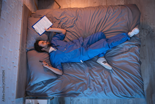 Young sleepwalker in doctor uniform holding pen and clipboard while sleeping on bed photo