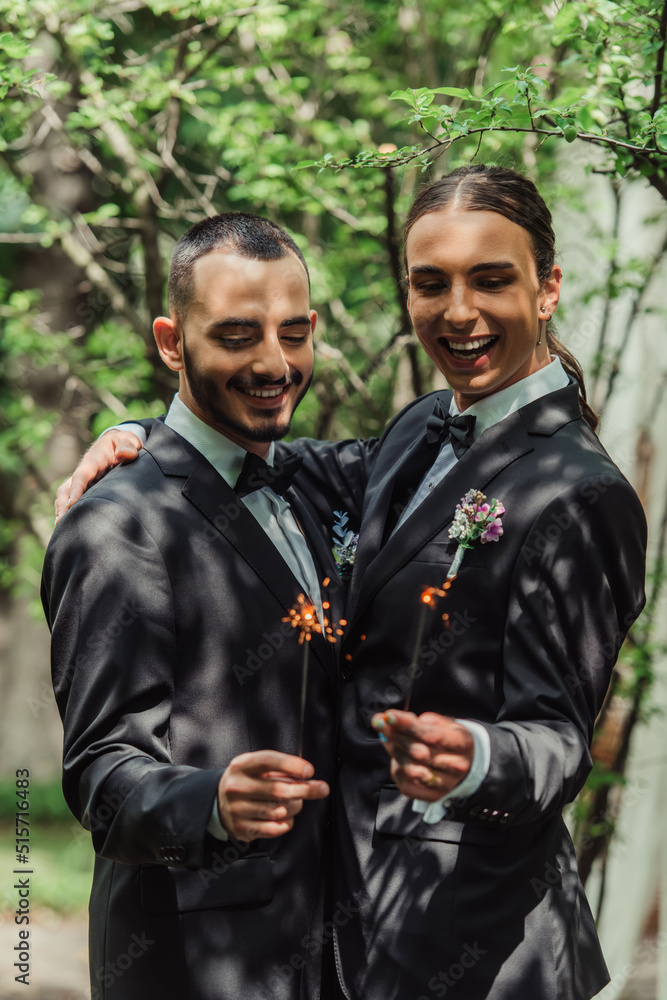 cheerful gay newlyweds in formal wear with boutonnieres holding sparklers in green park.