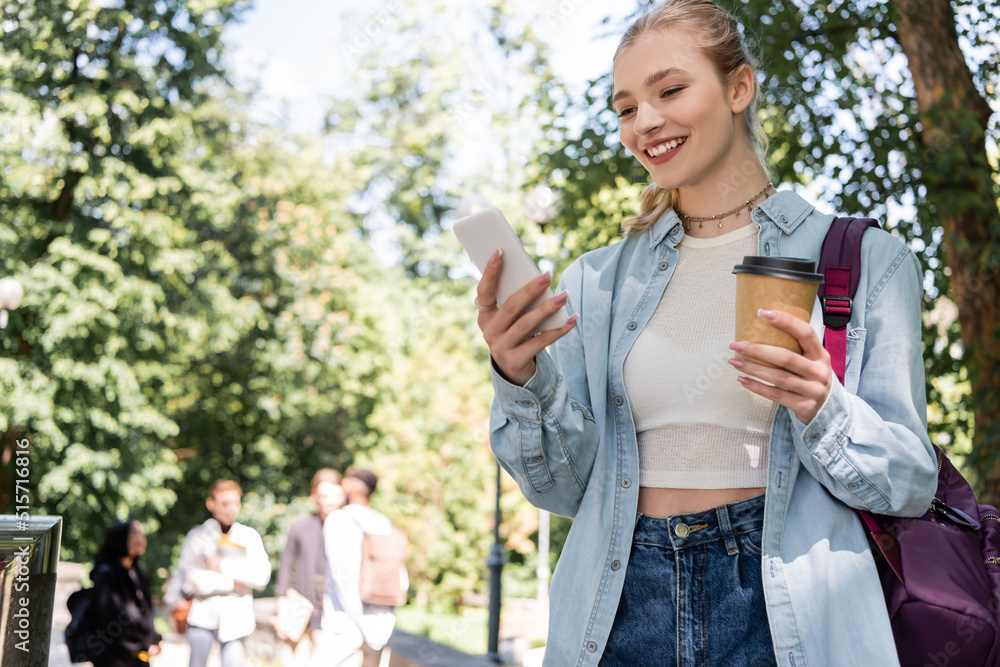 Smiling student using smartphone and holding coffee to go in park. Smiling student using smartphone and holding coffee to go in park.