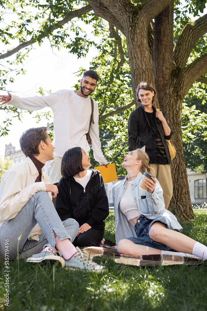 Positive interracial students with backpacks looking at friends on blanket in park. Positive interracial students with backpacks looking at friends on blanket in park.