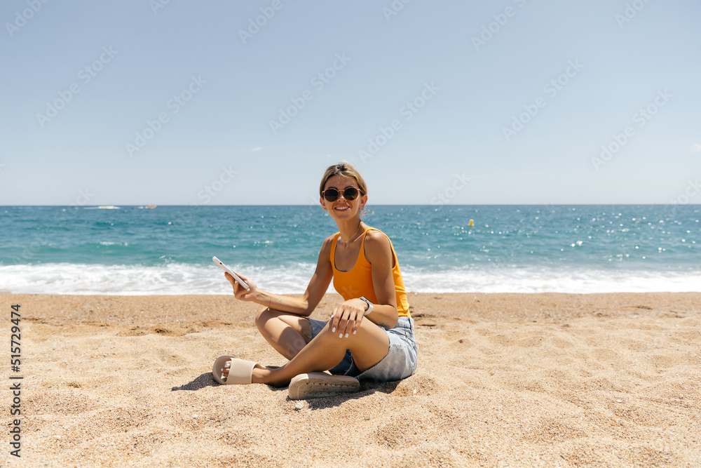 Full-lenght outdoor photo of smiling attractive woman with collected hair and tanned skin sitting on the beach with phone. Adorable girl using photo on the beach in sunlight 