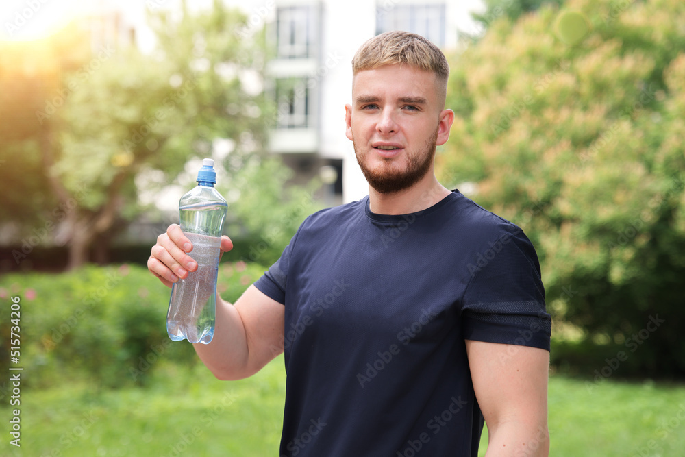 Portrait of handsome guy, young happy thirsty man runner is drinking pure fresh water from plastic bottle at warm sunny summer day outdoors after running, jogging, training outside, looking at camera Portrait of handsome guy, young happy thirsty man runner is drinking pure fresh water from plastic bottle at warm sunny summer day outdoors after running, jogging, training outside, looking at camera