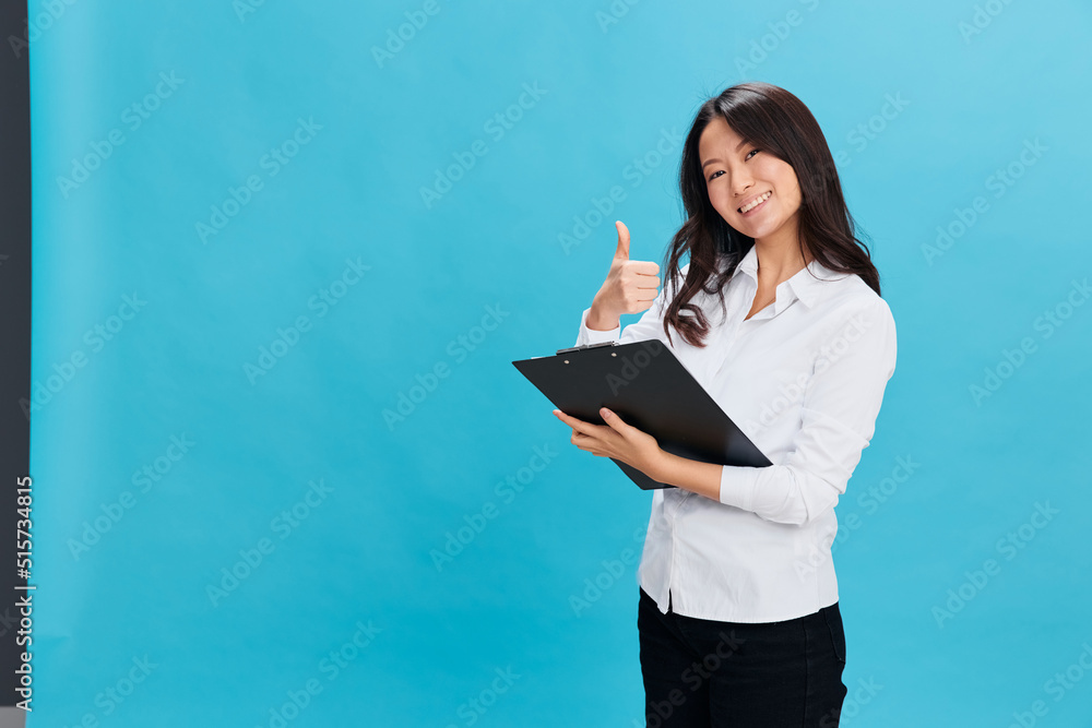 Smiling cute Asian businesswoman in classic dress code holds folder tablet with application form show thumb up gesture posing isolated on blue background. Cool business offer. Job interview concept Smiling cute Asian businesswoman in classic dress code holds folder tablet with application form show thumb up gesture posing isolated on blue background. Cool business offer. Job interview concept