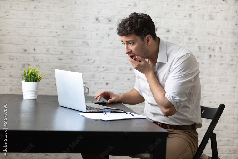 Shocked businessman after reading bad news in laptop about his business sits at the desk with documents. Crisis at work and stress concept Shocked businessman after reading bad news in laptop about his business sits at the desk with documents. Crisis at work and stress concept