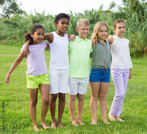 Portrait of five smiling kids who are hugging and posing in a park at summer day
