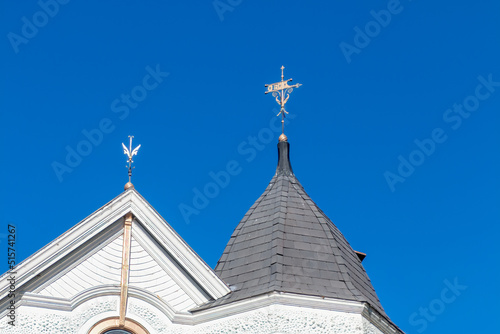 A weathervane is sitting on top of a dome and a decorative lighting rod is on a peaked roof. The weathervane an FS vane and the lighting rod is decorative. Both are metal. A blue sky is in back.