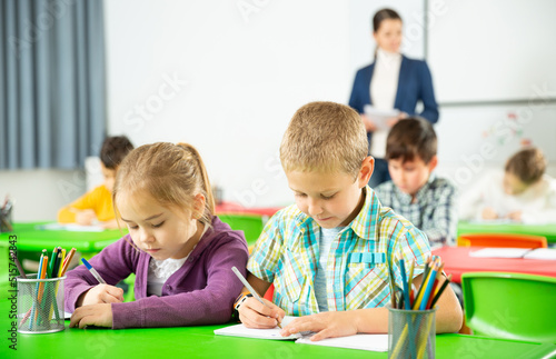 Assiduous school kids with pens and notebooks studying in classroom with female teacher