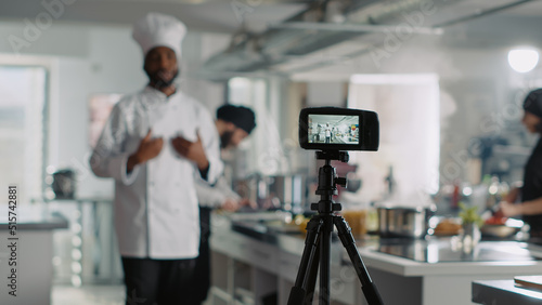 Vlogging camera filming male cook on television cooking show, talking about gastronomy and cuisine. Gourmet chef in uniform giving online food lesson, recording video on camera. Tripod shot.