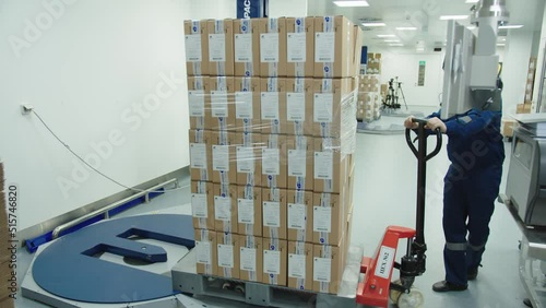 Loader in protective mask and uniform operates manually the cargo trolley at the pharmaceutical plant. factory worker transports the batch of packaged boxes of drugs.
