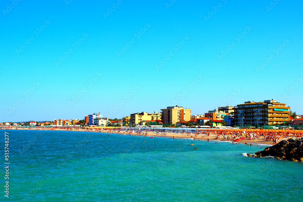 Beautiful overview of the Fano seaside with numerous bathers enjoying the crystal clear azure waters, stones on the right and buildings in the background on a sunny summer day