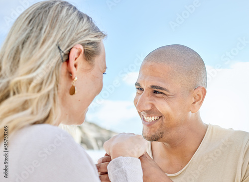A young man proposing to his girlfriend at the beach in summer. Happy interracial couple holding hands and smiling. Soon to be husband and wife staring lovingly into one anothers eyes outside