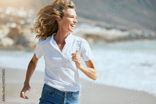 Cheerful mature woman running on the beach on a sunny day. Beautiful middle aged woman laughing, being active and having fun during summer vacation. Energetic lady spending her free time by the sea #515754433