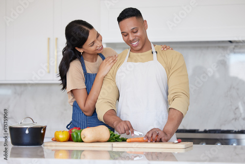 A young loving latin couple cooking a healthy dinner together at home. A loving man cutting carrots and other vegetable foods, preparing a meal and his girlfriend holds him and smiles in the kitchen