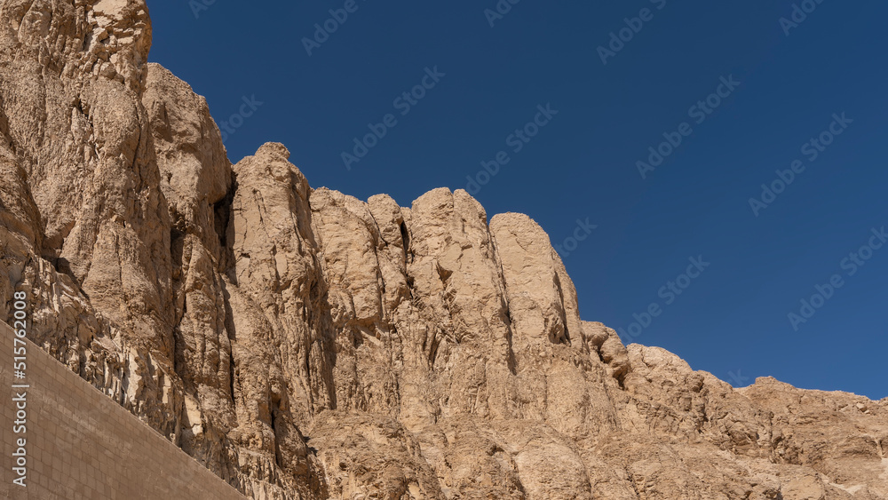 A picturesque cliff with steep slopes, devoid of vegetation, against a clear blue sky. A fragment of the wall of the burial temple of Queen Hatshepsut is visible. Egypt. Luxor. Copy Space