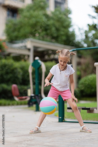 Candid lifestyle portrait of happy Cheerful caucasian little girl with blonde hair seven years old holding game ball for basketball outdoor at hot summer day